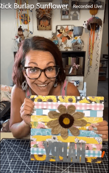 Amanda holding the completed DIY project of a Burlap Sunflower with sunflower themed scrapbook paper on paint sticks and the word happy underneath.