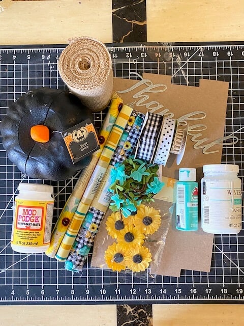 Supplies needed to make a door hanger from a dollar tree foam pumpkin, sunflower and teal plaid fabric bow, burlap, and a galvanized metal "thankful".