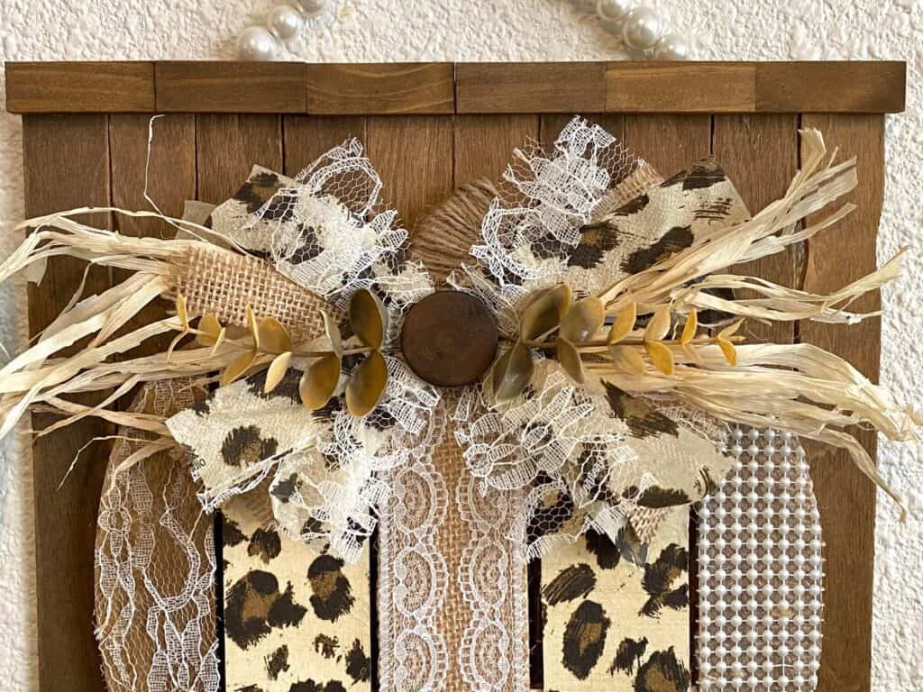 Top of the dollar Tree pumpkin with a messy bow with lace, burlap, leopard print napkin, faux eucalyptus, and a brown button with the stem of the pumpkin wrapped in twine.