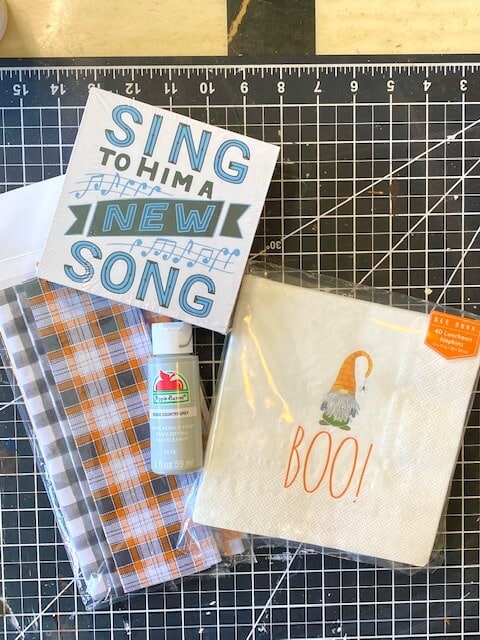 Supplies needed to make a a shelf sitter box with a Rae Dunn Gnome napkin and orange and grey plaid fabric.