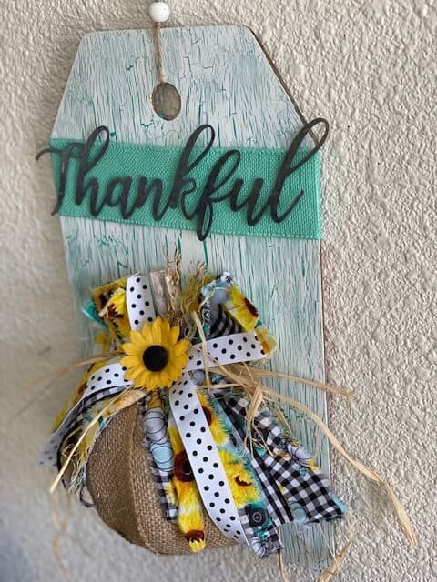 Dollar Tree Burlap Foam Pumpkin Thankful Door Hanger with Teal crackle paint background and sunflowers, truck, buffalo check bow and a wood bead hanger.