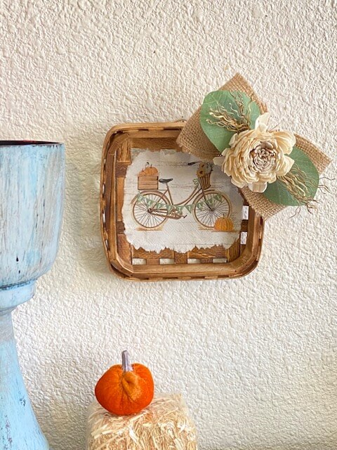 Fall Vintage Bicycle Tobacco Basket made with a cocktail napkin mod podged to the center of a small burlap bow with wood flower and lambs ear, next to a velvet pumpkin.