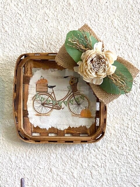 Fall Vintage Bicycle Tobacco Basket made with a cocktail napkin mod podged to the center of a small burlap bow with wood flower and lambs ear, hanging on a white wall.