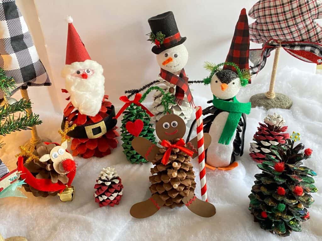 Pine Cone Santa Claus Kids Christmas Crafts with pine cone snowman, penguin, gingerbread man, rudolph the red nosed reindeer, and a christmas tree on faux snow.
