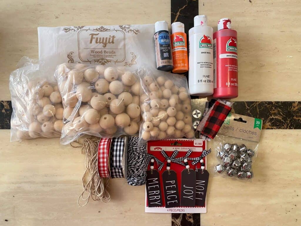 Supplies needed to make a snowman woodbead garland with black, white, and red painted beads, "noel" Christmas ornament, twine tassel, silver bells.