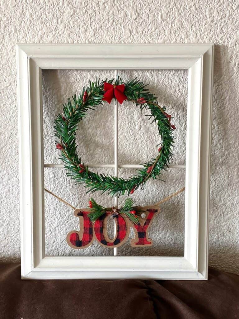 White thrift store frame with a faux pine wreath and a red bow and an ornament that says "joy".