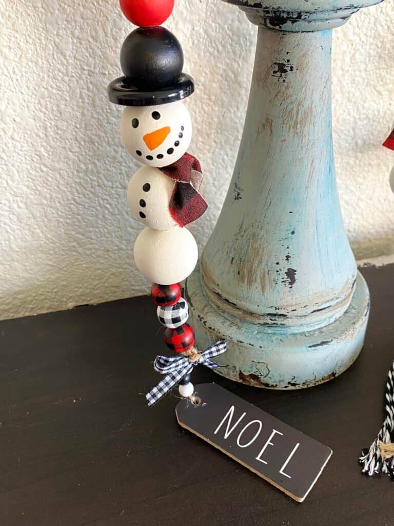 Snowman with a black top hat made out of wood beads with decorative buffalo check small beads on the end with a "noel" tag ornament.