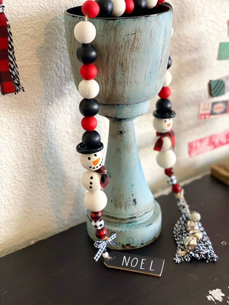 Snowman with a black top hat made out of wood beads with decorative buffalo check small beads on the end with a "noel" tag ornament.