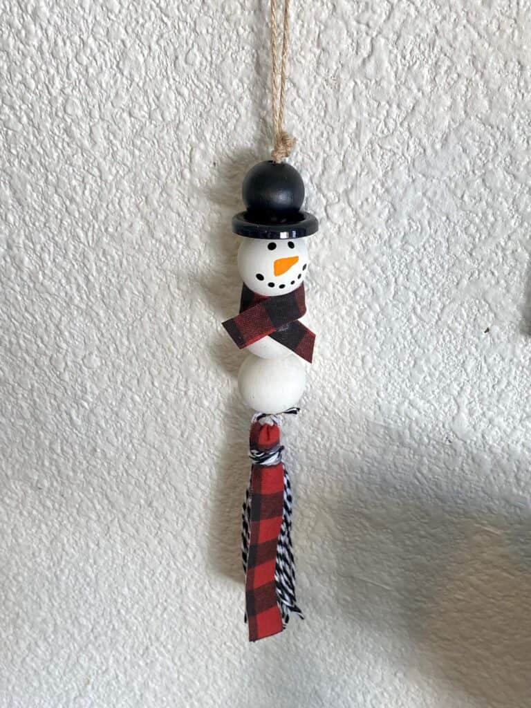 Snowman with a black top hat made out of wood beads with decorative red and black buffalo check ribbon and bakers twine tassel.