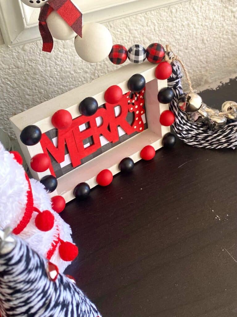 Looking down on a bookshelf with a "Merry" wooden frame shelf sitter with buffalo check and red and lack half wood beads.