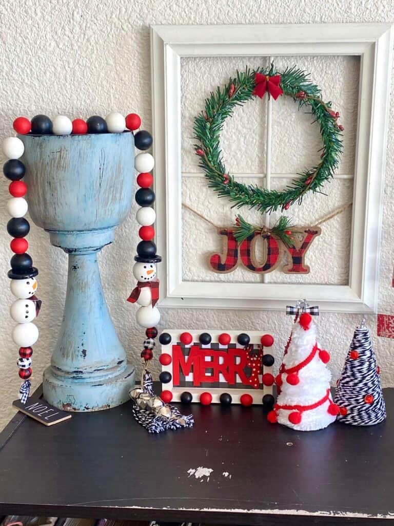 Display of black white and red DIY christmas crafts and decor with a shalf sitter, foam cone christmas trees, a wreath window frame and a snowman wood bead garland.