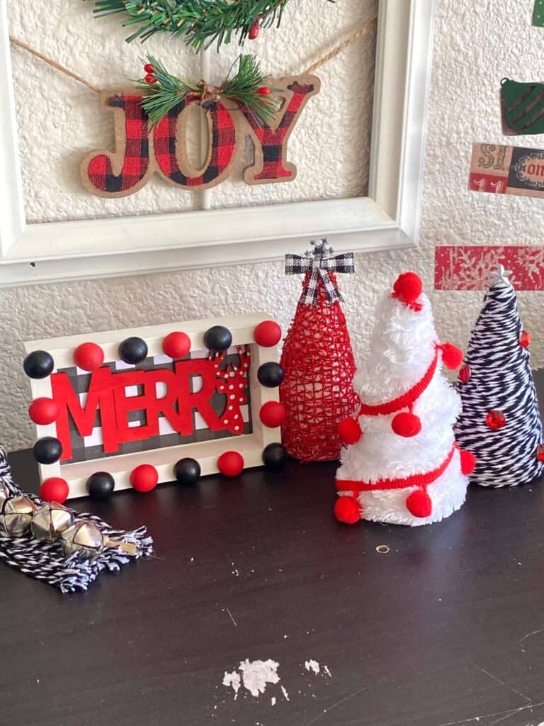 Red, white, and black themed DIY Christmas decor on a bookshelf. 3 foam cone christmas trees and a "Merry" black white buffalo check shelf sitter.
