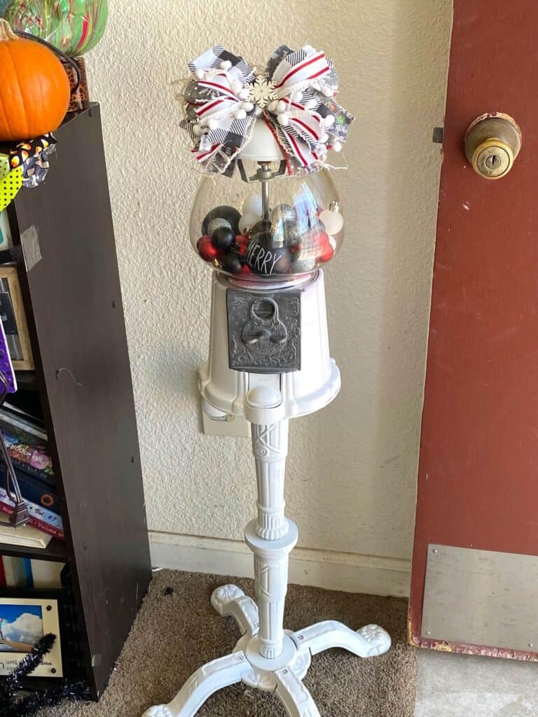 Christmas decorated thrift store vintage gumball machine that is painted white, with red, black, silver, and white ornament bulbs inside and a big messy bow on top.