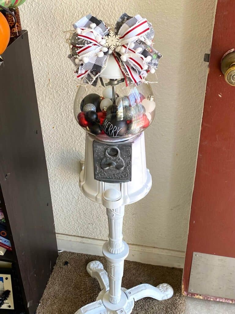 Christmas decorated thrift store vintage gumball machine that is painted white, with red, black, silver, and white ornament bulbs inside and a big messy bow on top.
