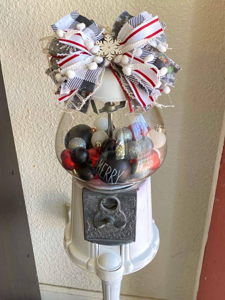 Christmas decorated thrift store vintage gumball machine that is painted white, with red, black, silver, and white ornament bulbs inside and a big messy bow on top.