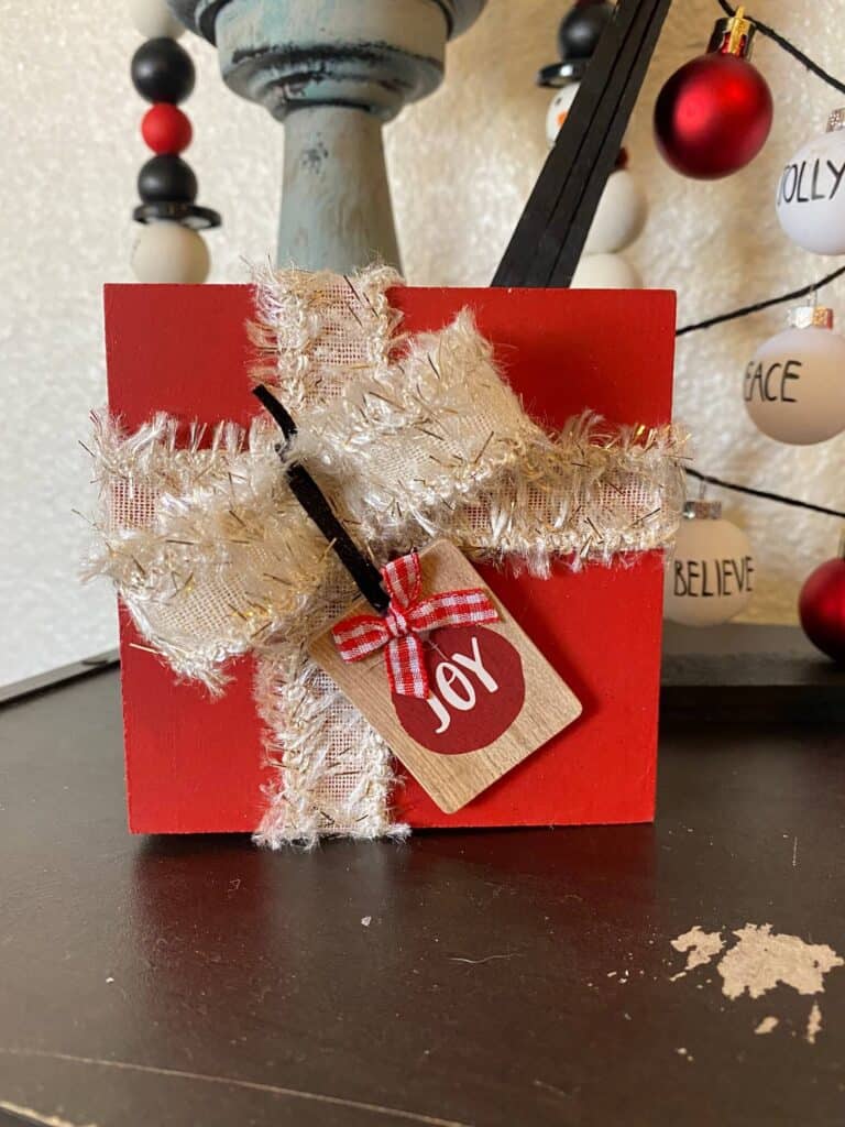 Red wood block present with white and old ribbon and a mini "joy" ornament.