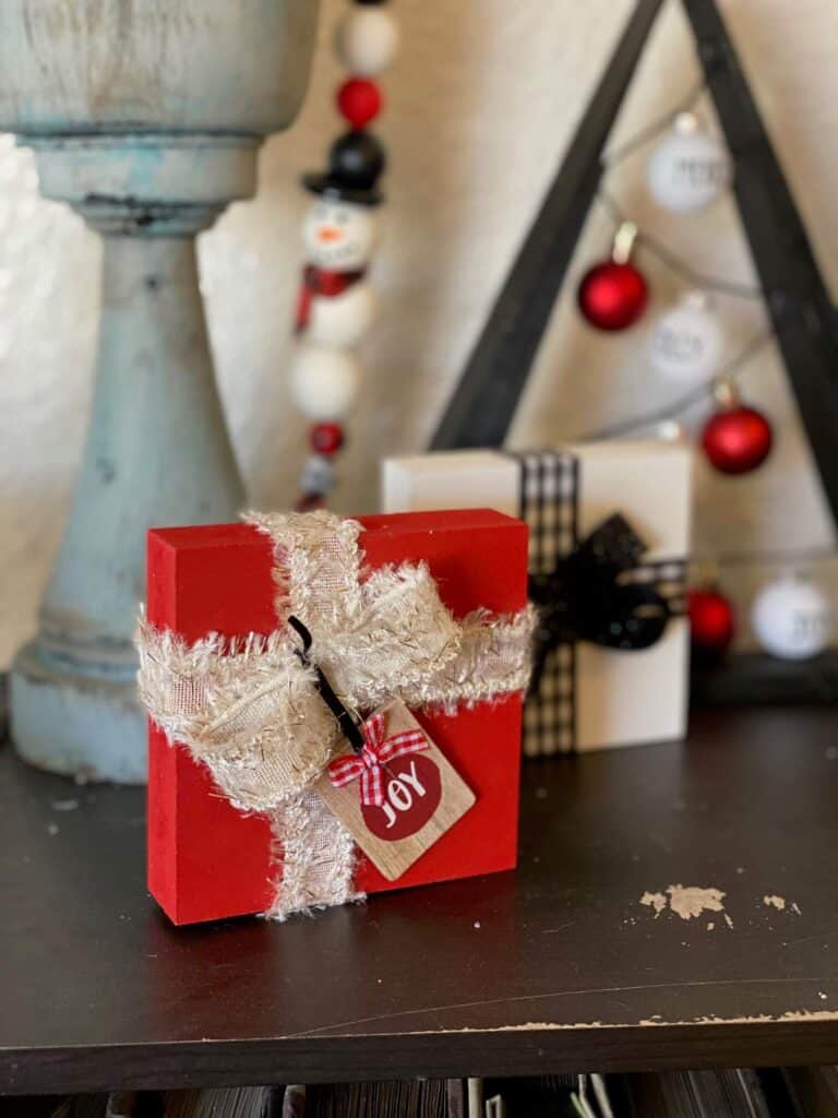 Red wood block present with white and old ribbon and a mini "joy" ornament.