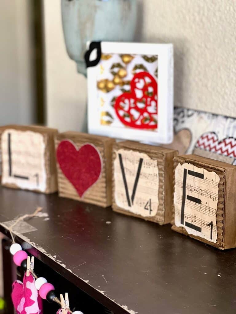 Love blocks with a red glitter heart for the "O" and corrugated cardboard with music sheet scrapbook paper with burnt edges on top. the letters are laid out horizontally.