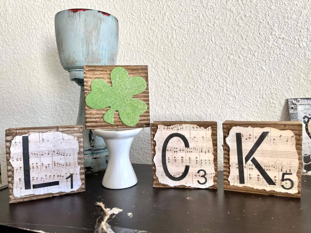 4 square blocks that spell the word "luck" and the "U" is a green glitter shamrock. Blocks are covered in corrugated cardboard with music sheet scrapbook paper with burnt edges on top. The "U"