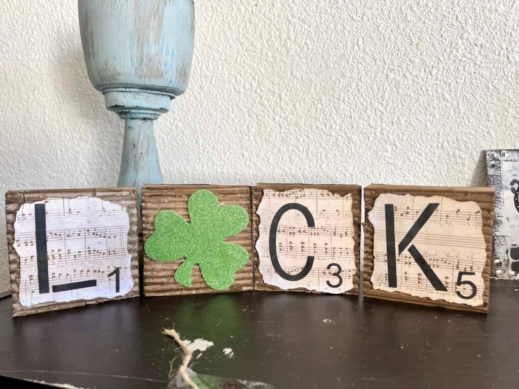 4 square blocks that spell the word "luck" and the "U" is a green glitter shamrock. covered in corrugated cardboard with music sheet scrapbook paper with burnt edges on top. Letters laid out horizontal.