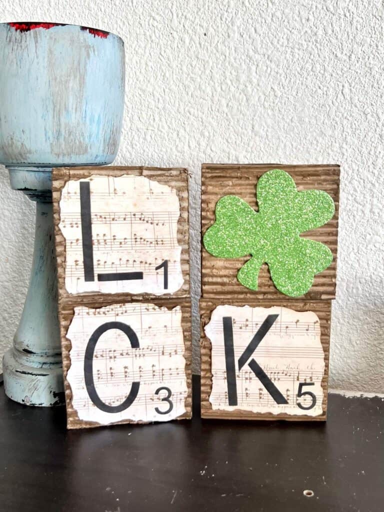 4 square blocks that spell the word "luck" and the "U" is a green glitter shamrock. covered in corrugated cardboard with music sheet scrapbook paper with burnt edges on top.