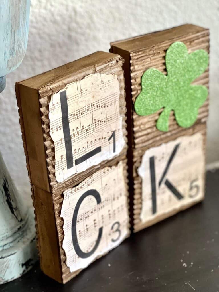 4 square blocks that spell the word "luck" and the "U" is a green glitter shamrock. covered in corrugated cardboard with music sheet scrapbook paper with burnt edges on top, the blocks are stacked on top of each other, 2 on top and 2 on bottom.