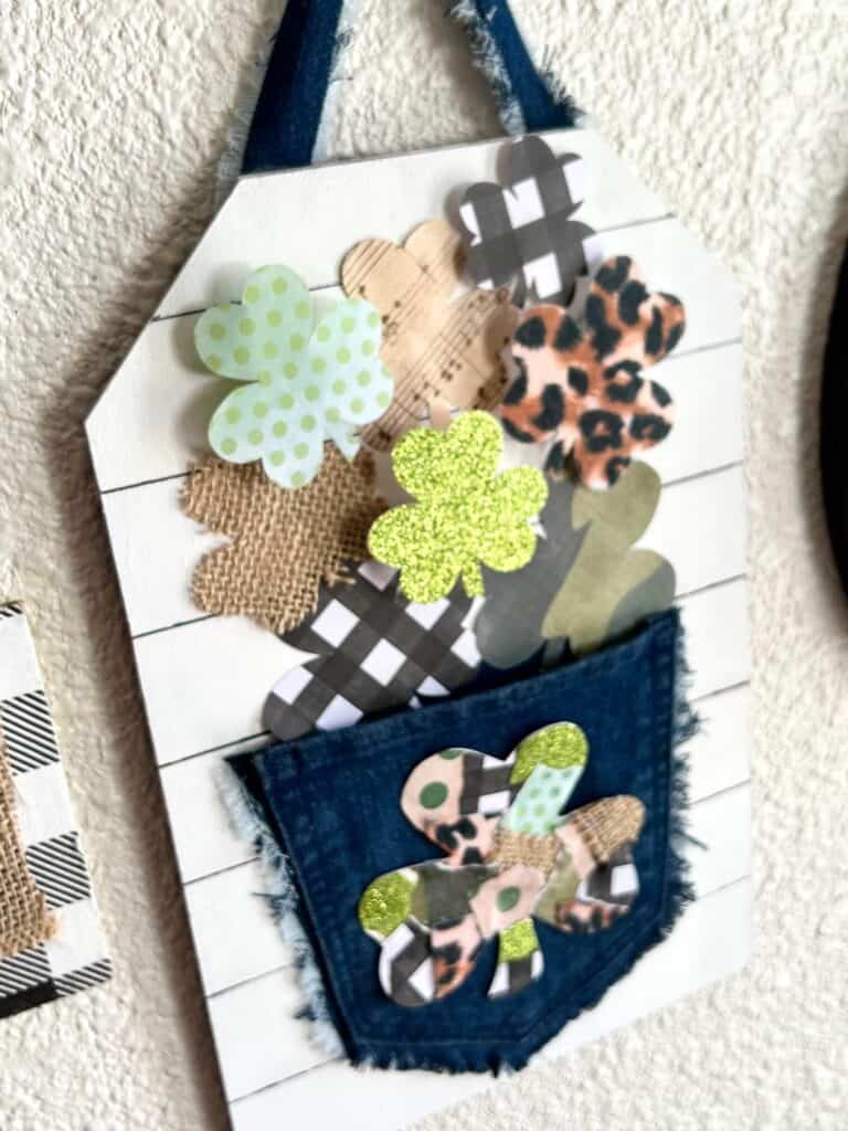 Wild Patchwork Shamrocks spilling out of a jeans pocket that are green glitter, leopard print, buffalo check, music sheet, and burlap, and camo with a faux shiplap background for DIY St. Patrick's Day and St. Patty's crafts and decor.