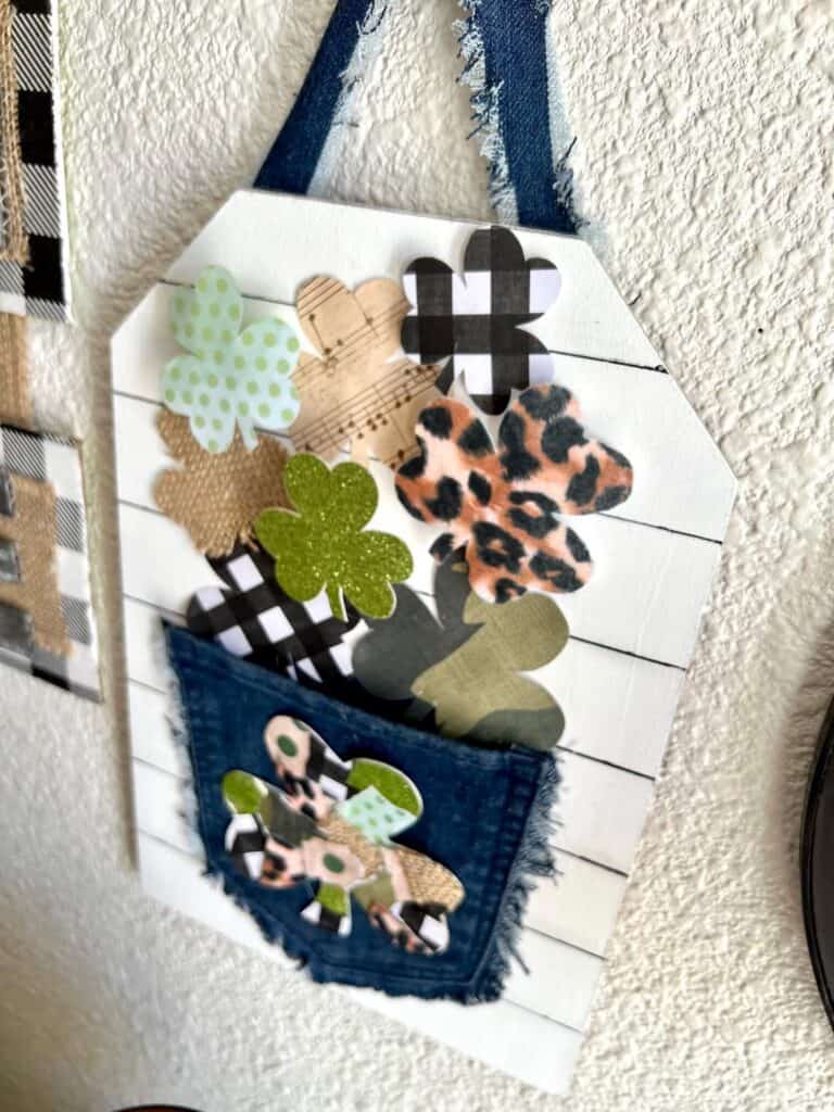 Patchwork Shamrock on the back of a jeans pocket with smaller shamrocks spilling out of the pocket that are green glitter, leopard print, buffalo check, music sheet, and burlap with a faux shiplap background.