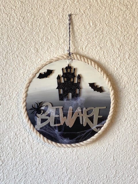 Dollar Tree DIY Halloween Beware sign for your front door with a haunted house, bats, and fake cobwebs, and nautical rope around the edge of the round wood sign.