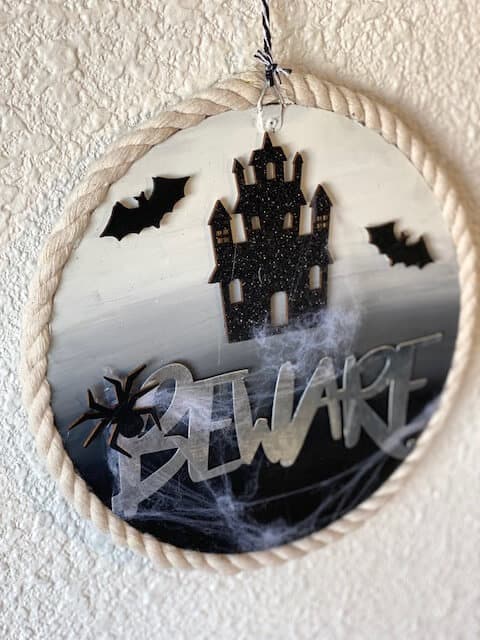 Dollar Tree DIY Halloween Beware sign for your front door with a haunted house, bats, and fake cobwebs, and nautical rope around the edge of the round wood sign.