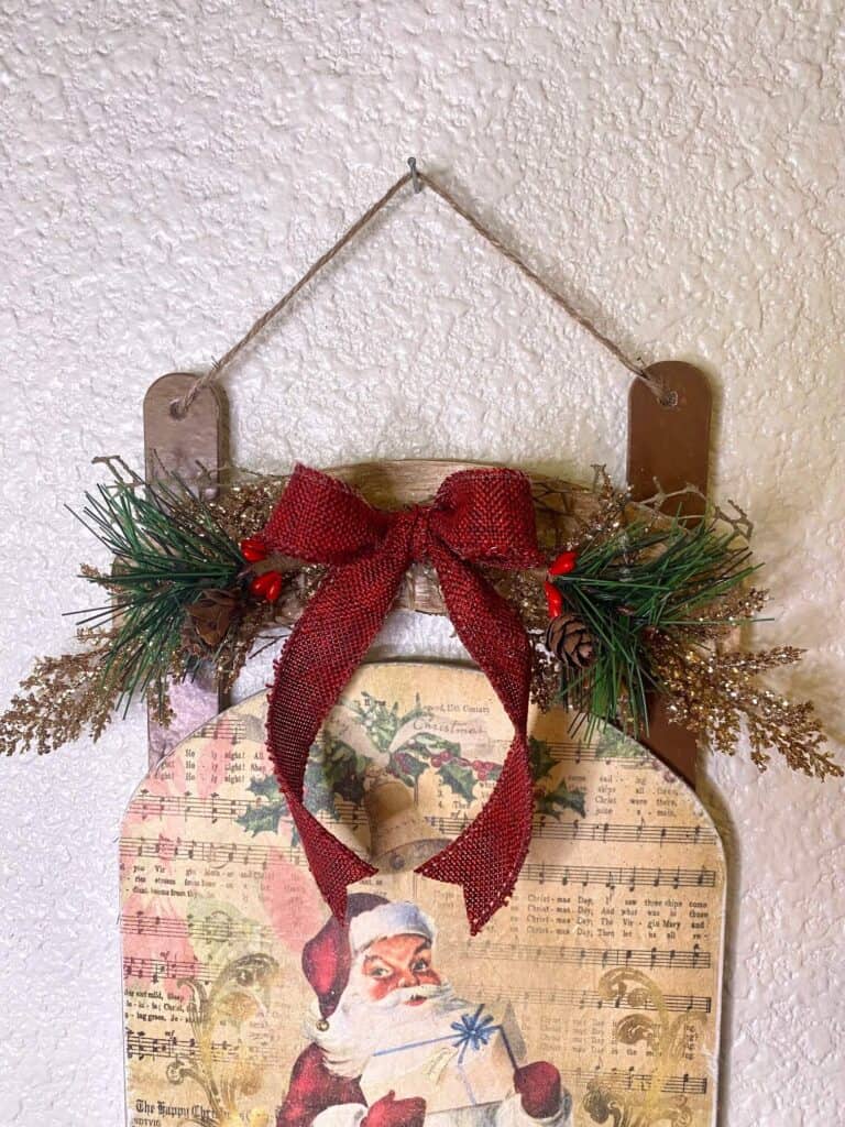 The top of the sleigh makeover with gold leaf picks holly berry picks, and a deep red burlap shoelace bow with a simple twine hanger.