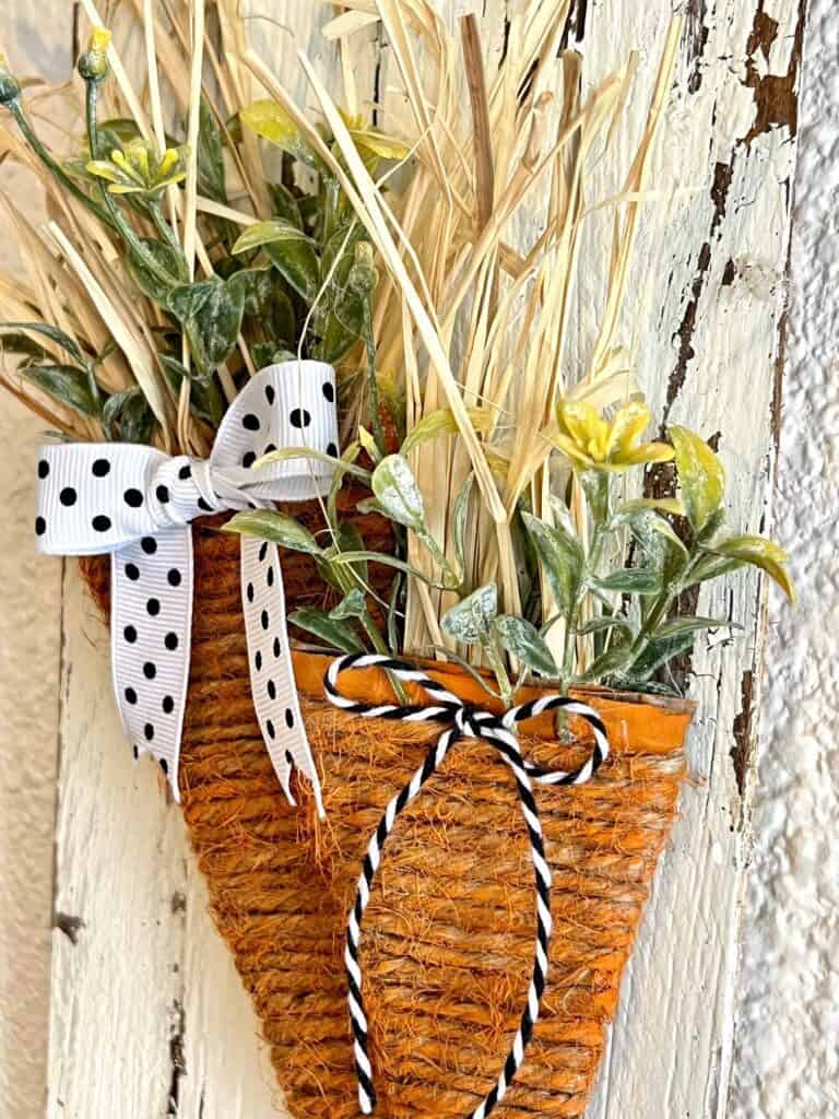 Close up of the raffia and greenery coming out the top of the twine wrapped cardboard carrots with small black and white bows.