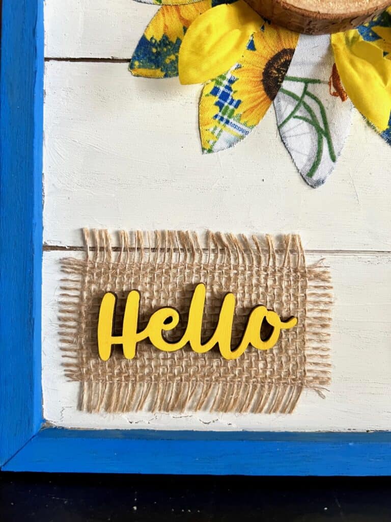 Wooden cutout of the word "Hello" painted yellow on a piece of burlap background.