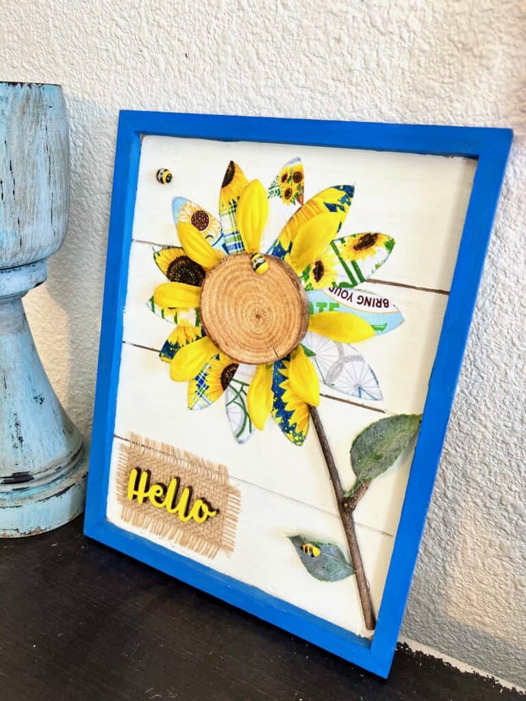 DIY Spring or Summer Fabric Sunflower Decor craft project made with blue and yellow theme sunflower fabric a mini bees and a wooden hello with burlap, in a blue frame. 