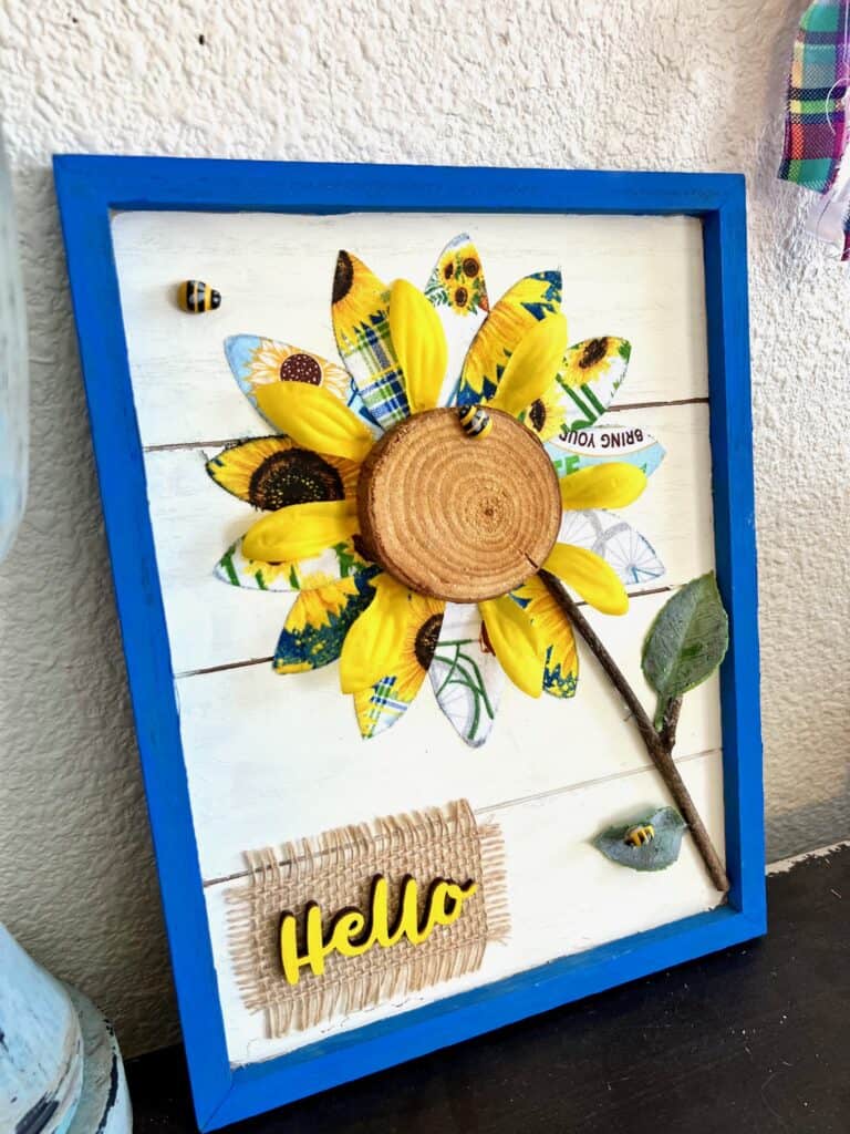 DIY Spring or Summer Fabric Sunflower Decor craft project made with blue and yellow theme sunflower fabric a mini bees and a wooden hello with burlap, in a blue frame. 