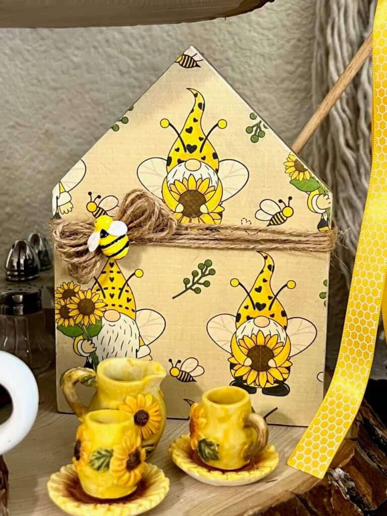 Front of the Dollar Tree wooden house with sunflower gnome free printable craft paper and a mini bee on top of twine decor bow, with a mini sunflower tea set in front.