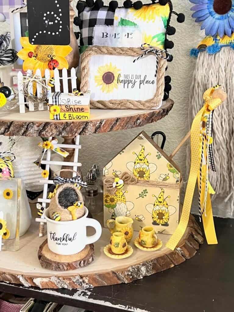 Bottom tier of the rustic wooden 3 tiered tray showing the sunflower gnome house along with the mini tea set and a mini mug that says "thankful for you" with a small twine bee hive.