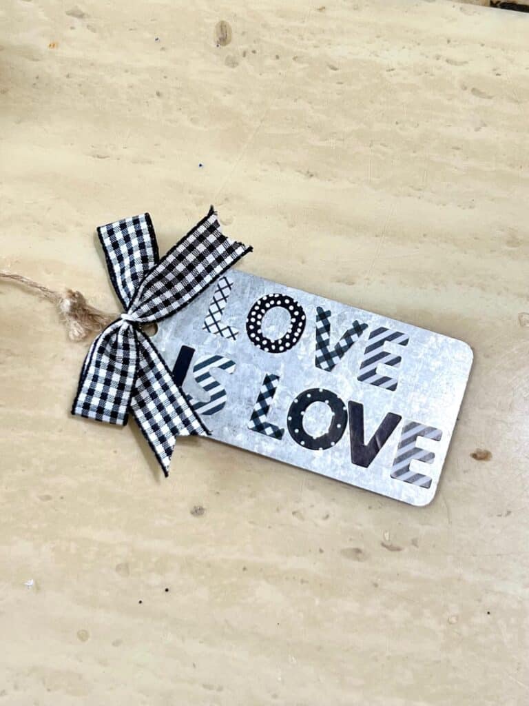 Galvanized metal hang tag with stickers that say  "love is love" and a small buffalo check bow. attached to the end of the beaded garland.