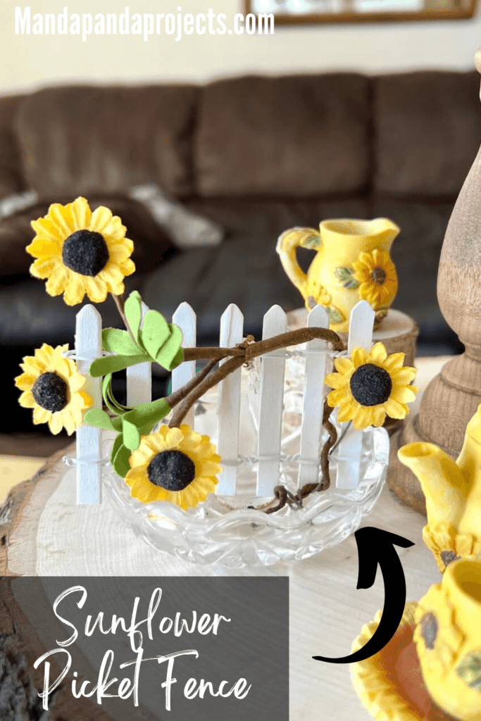 Small white picket fence with sunflowers on a vine wrapped around the fence, in a glass dish.