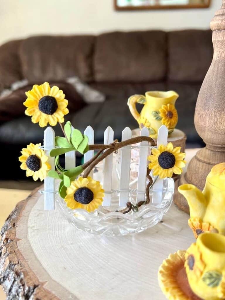 Small white picket fence with sunflowers on a vine wrapped around the fence, in a glass dish.