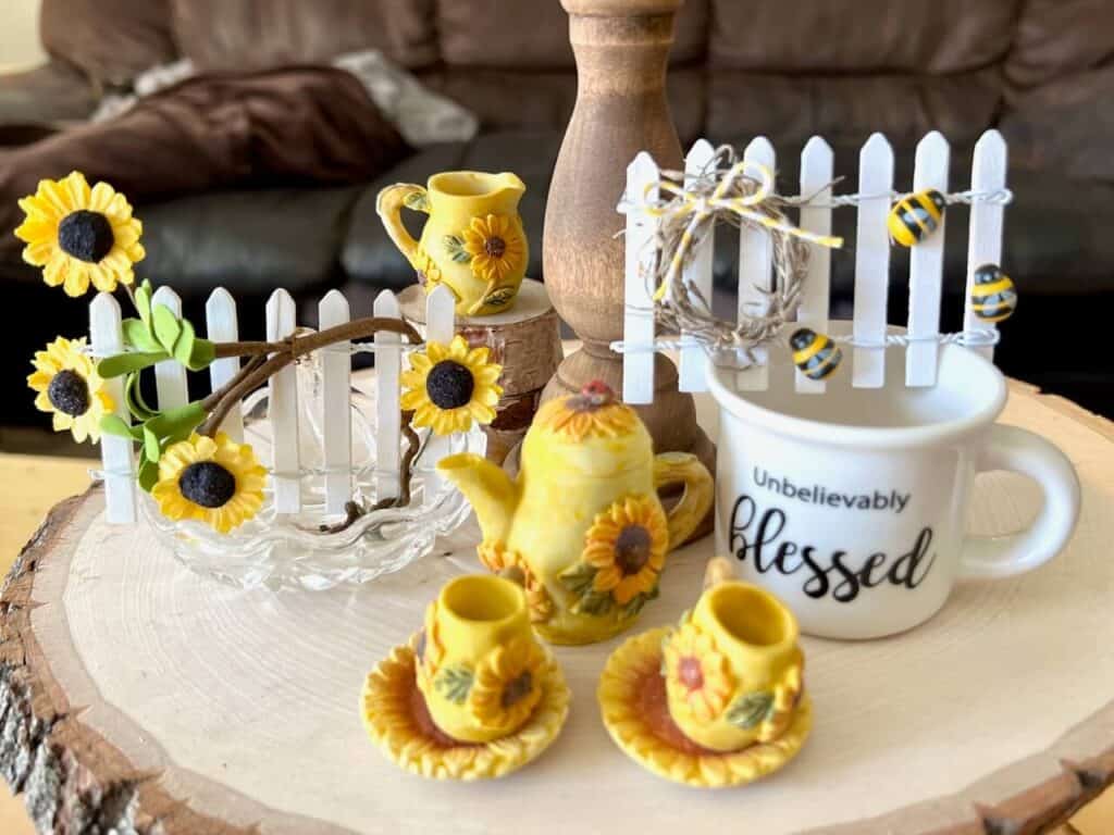 Sunflower and bee Tiered Tray Mini Picket Fence. 2 little white fences, one with a sunflower vine and one with a mini wreath and bumblebees, sitting on a wooden tray with a mini tea set and mini mug.