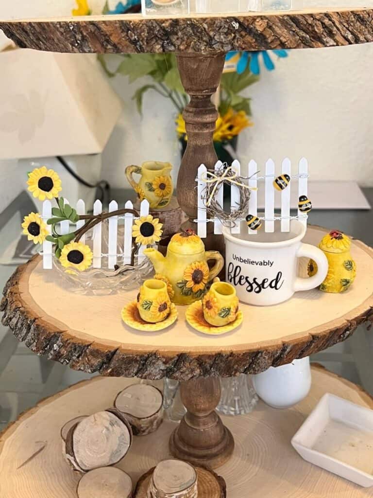 Sunflower and bee Tiered Tray Mini Picket Fence. 2 little white fences, one with a sunflower vine and one with a mini wreath and bumblebees, sitting on a wooden tray with a mini tea set and mini mug.