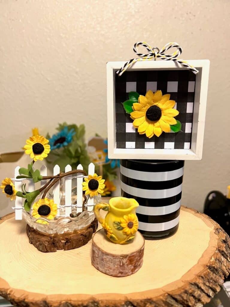 White sunflower picket fence on a wooden round next to a mini sunflower tea pot and a buffalo check sunflower decor.