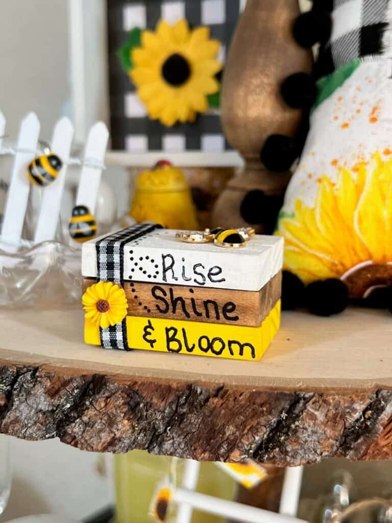 Front of the mini bookstack decor showing the words Rise Shine & Bloom, with buffalo check ribbon and a mini sunflower.