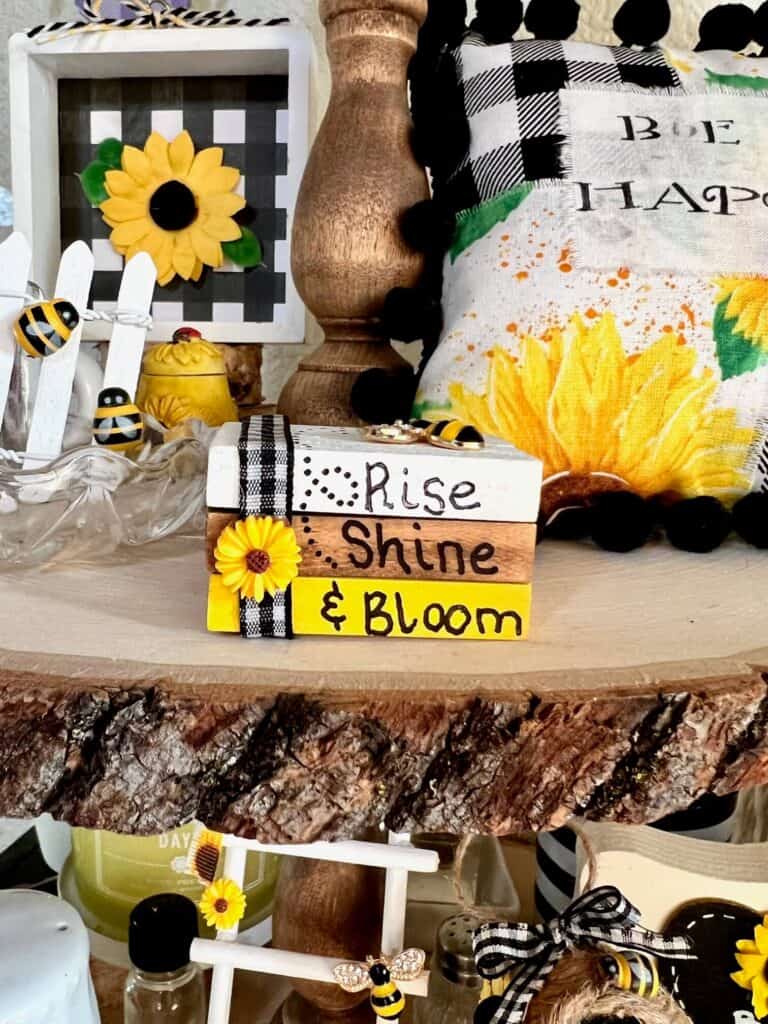 Front of the mini bookstack decor showing the words Rise Shine & Bloom, with buffalo check ribbon and a mini sunflower with a decorative pillow in the background.
