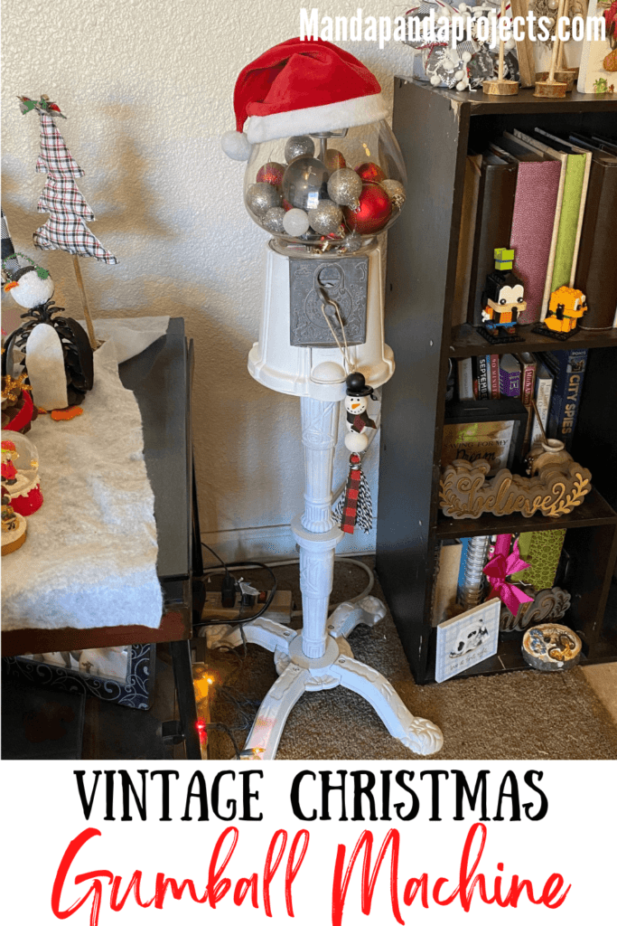 Christmas decorated thrift store vintage gumball machine that is painted white, with red, black, silver, and white ornament bulbs inside, a santa hat sitting on top, and a snowman wood bead garland ornament hanging from the coin turner with DIY tassel.