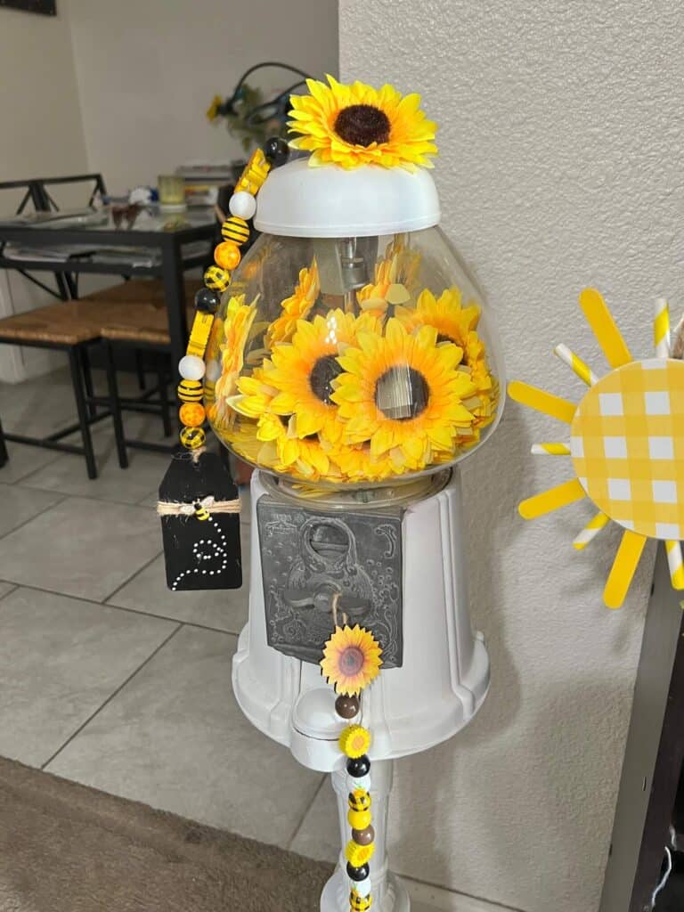 Sunflower Vintage Gumball Machine with sunflower heads inside and on top, and sunflower and bee mini wood bead garlands hanging form the top and coin turner. Gumball machine is painted all white.