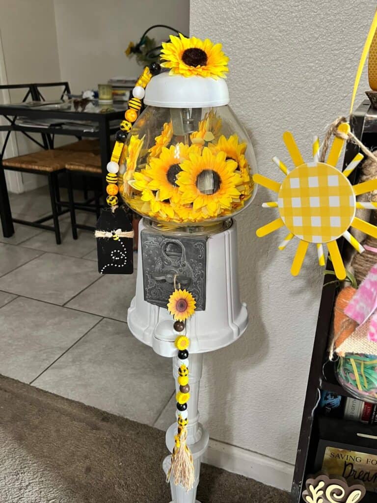 Sunflower Vintage Gumball Machine with sunflower heads inside and on top, and sunflower and bee mini wood bead garlands hanging form the top and coin turner. Gumball machine is painted all white.