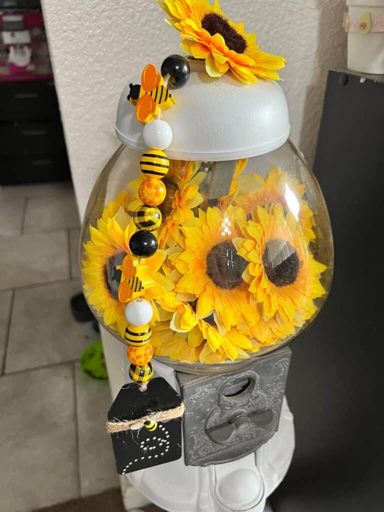 Top portion of the vintage gumball machine filled with sunflower heads and a sunflower on the top with a Bumblebee wood bead garland hanging off the side.