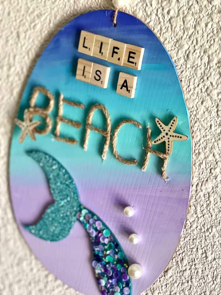 Dollar Tree Ombre Mermaid Tail DIY summer nautical beach decor with "Life is a Beach" written in scrabble tiles and jute twine and a pearl bead hanger, along with mermaid purple, blue and teal sequins and Ombre paint.